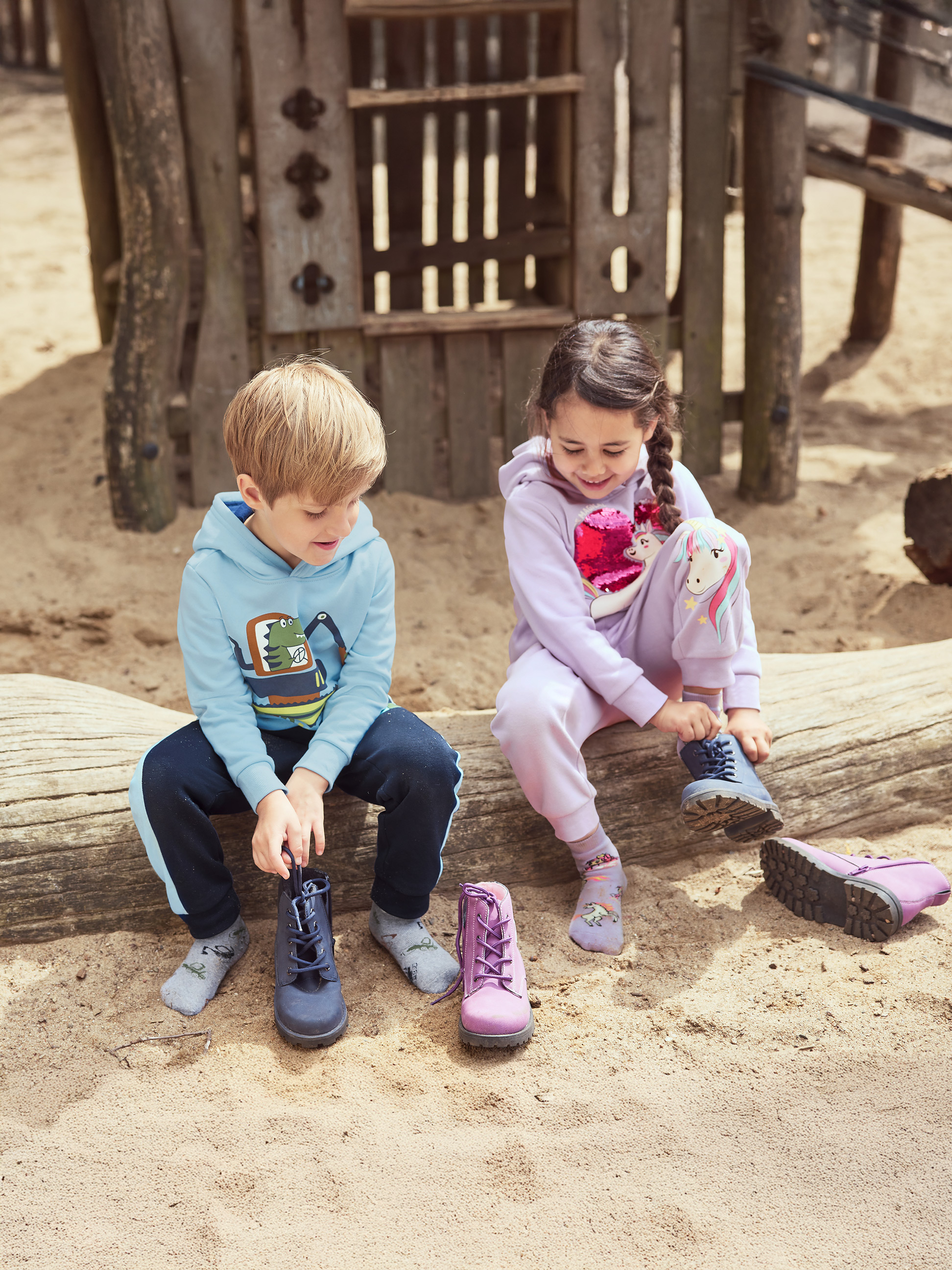 Deux enfants attachent leurs bottes en étant assis sur une bûche dans un bac à sable.