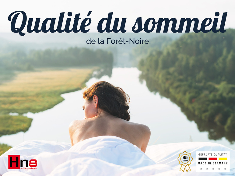 Une femme allongée sur un drap blanc avec vue sur un lac et le logo 'Hna Schlafsysteme' avec 'Geprüfte Qualität'