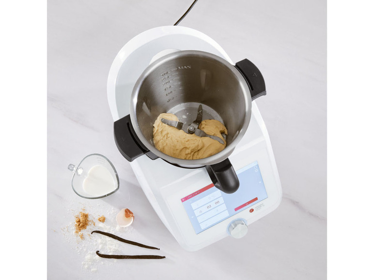 Un robot de cuisine blanc avec un bol de pâte et des ingrédients à côté.