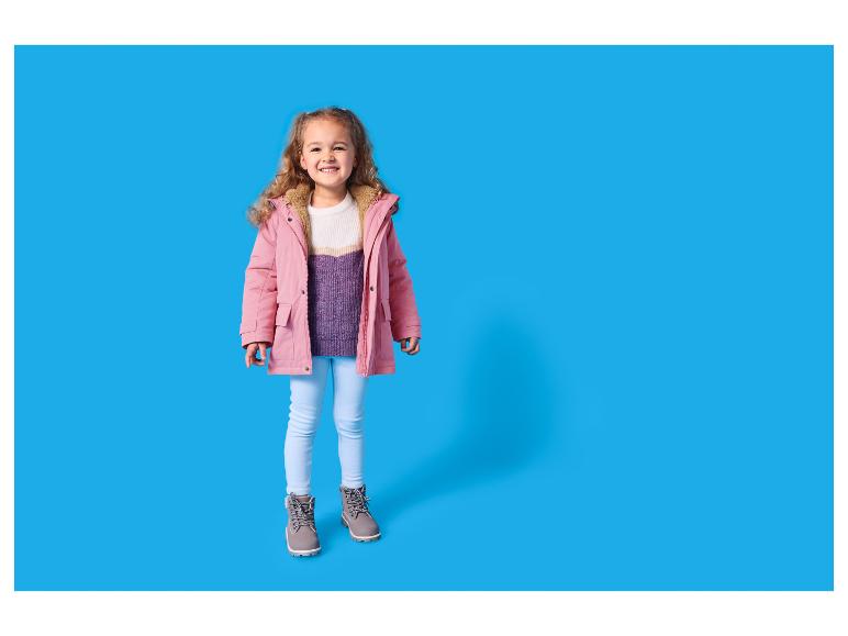 Fille souriante en veste d'hiver rose, pull blanc et violet, jean bleu clair et bottes grises.