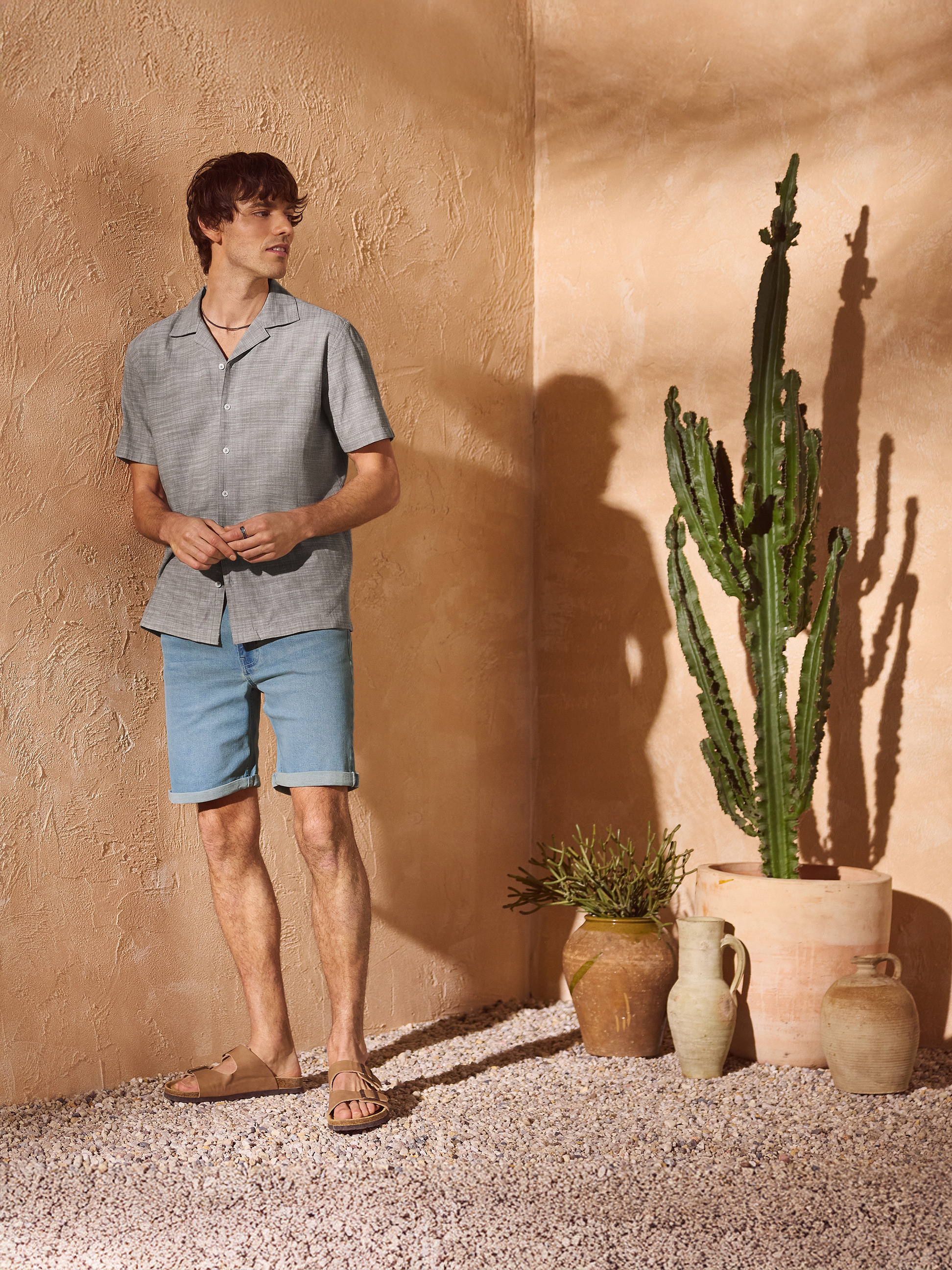 Homme en chemise grise et short en jean, sandales, dans une ambiance méditerranéenne.