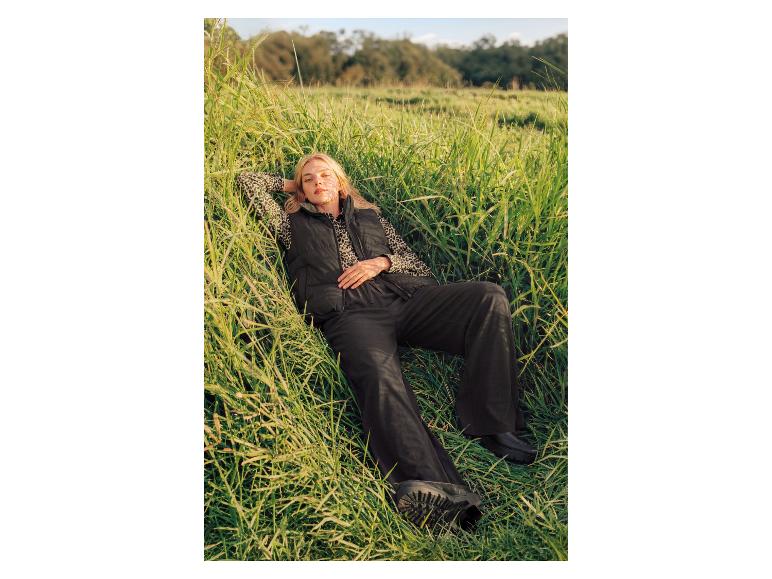 Femme en gilet matelassé noir, chemise imprimé léopard et pantalon noir allongée dans l'herbe haute.