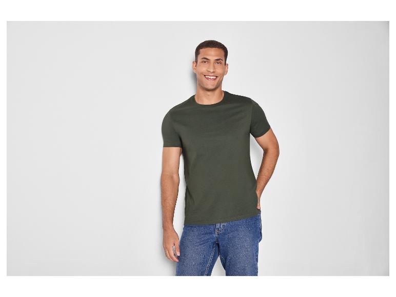 Homme en t-shirt vert foncé et jean bleu, souriant devant un mur blanc.