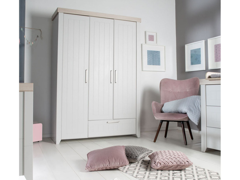 Une armoire blanche à trois portes et un tiroir, un fauteuil rose et des coussins décoratifs.