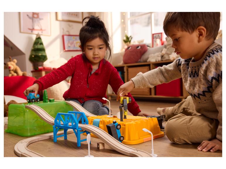 Deux enfants jouent avec un train en bois et des voitures jouets sur le sol.
