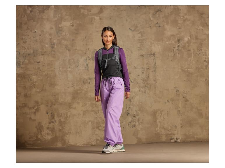 Une femme en pantalon violet, un gilet noir et des baskets devant un mur rugueux.