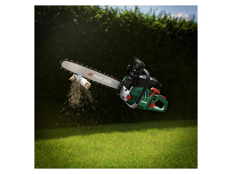 Tronçonneuse coupant une branche, avec de la sciure en l'air, sur une haie verte et de l'herbe.