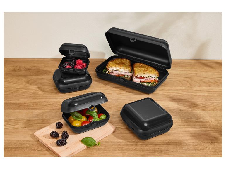 Boîtes à lunch noires avec sandwichs, baies et tomates sur une table en bois.