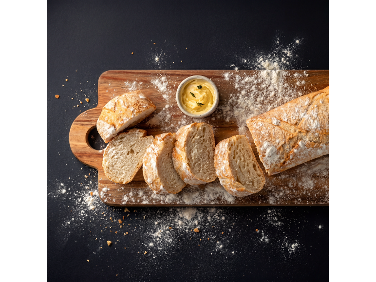 Pain ciabatta tranché sur une planche en bois avec du beurre et de la farine sur fond sombre.