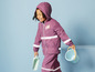 Un enfant porte un imperméable et un pantalon violets avec des bandes réfléchissantes et tient une pelle et un seau.