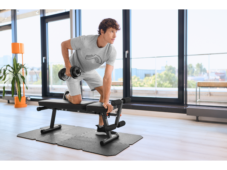 Homme s'entraînant avec un haltère et un banc de musculation sur un tapis d'exercice en intérieur.