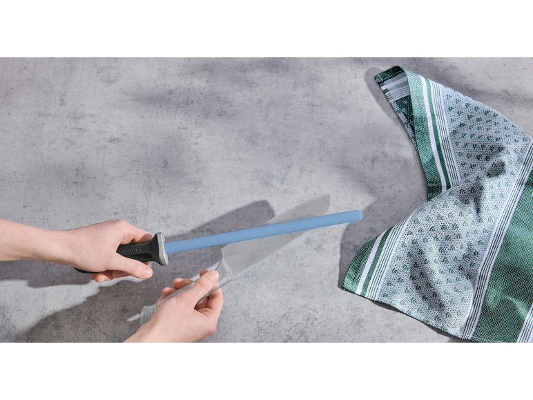 Mains aiguisant un couteau avec un fusil à aiguiser bleu, à côté d'un torchon vert et blanc.