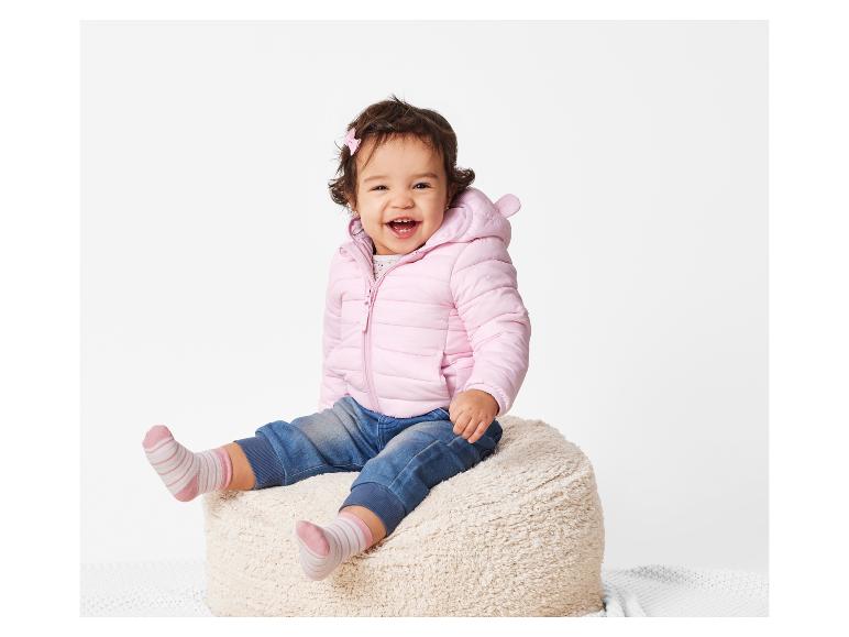 Un bébé souriant porte une veste rose et des chaussettes rayées assis sur un pouf.