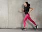 Femme en haut noir et leggings roses courant devant un mur en béton.