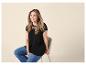 Femme souriante en t-shirt noir en dentelle et jean, assise sur un tabouret.