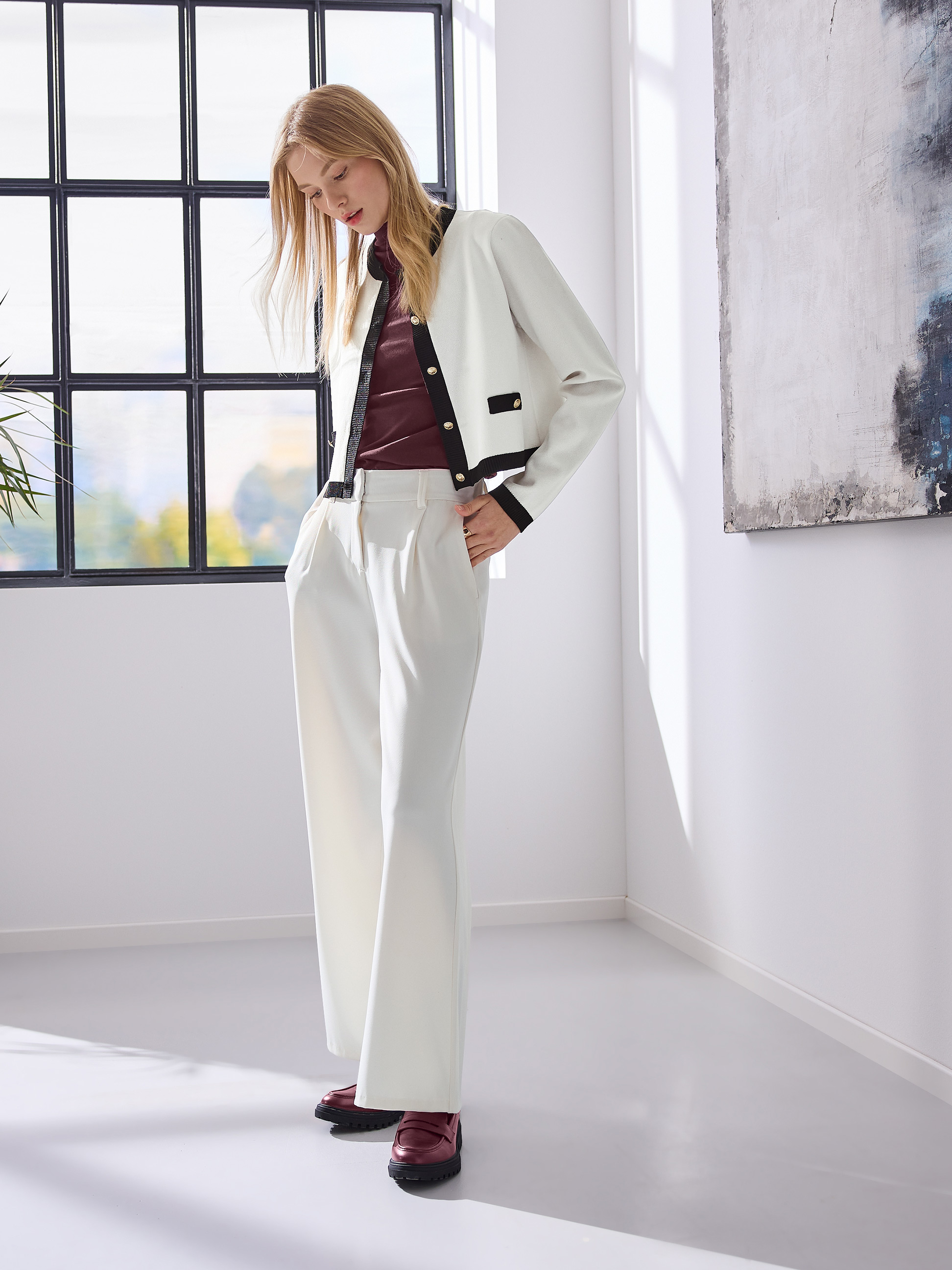 Femme en veste courte crème, pantalon large blanc et mocassins bordeaux, posant près d'une fenêtre.
