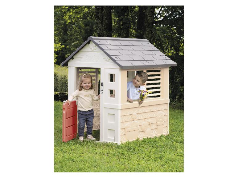 Deux enfants jouent dans une cabane en plastique avec un toit gris et une porte rouge dans un jardin.