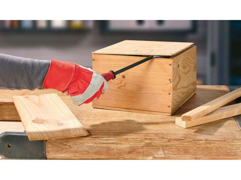 Une main portant un gant rouge ouvre une caisse en bois à l'aide d'un outil.