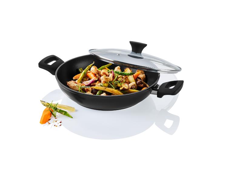Wok noir avec couvercle en verre, rempli de poulet et légumes sautés, avec des ingrédients frais à côté.