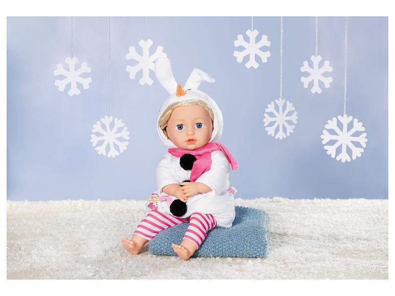 Poupée en tenue de bonhomme de neige avec écharpe rose, assise sur une couverture bleue parmi les flocons.