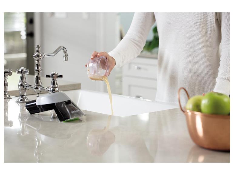 Une personne verse du liquide d'un récipient dans un évier de cuisine, avec des pommes vertes à proximité.