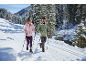 Couple en vestes et pantalons d'hiver avec bâtons sur une piste de ski en forêt enneigée.