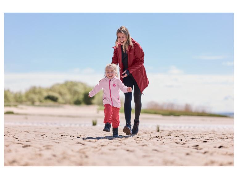 Une femme en manteau softshell outdoor rouge CRIVIT court sur la plage avec un enfant.