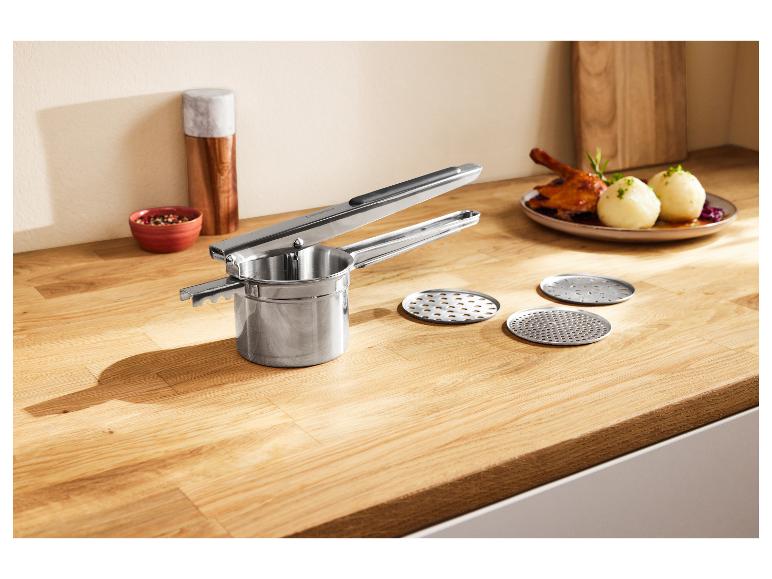 Un presse-purée en acier inoxydable avec des disques interchangeables sur un comptoir en bois, avec un dîner en arrière-plan.