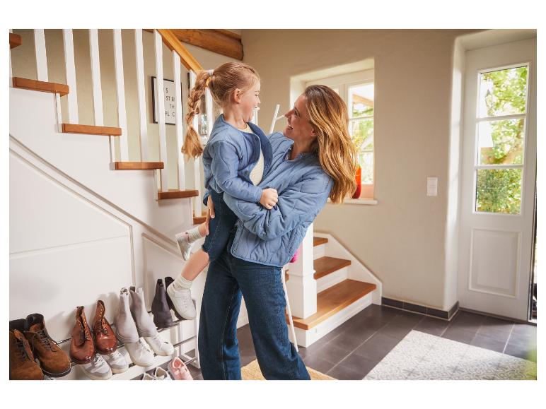 Une mère porte sa fille dans ses bras, elles sont devant un escalier avec une étagère à chaussures.