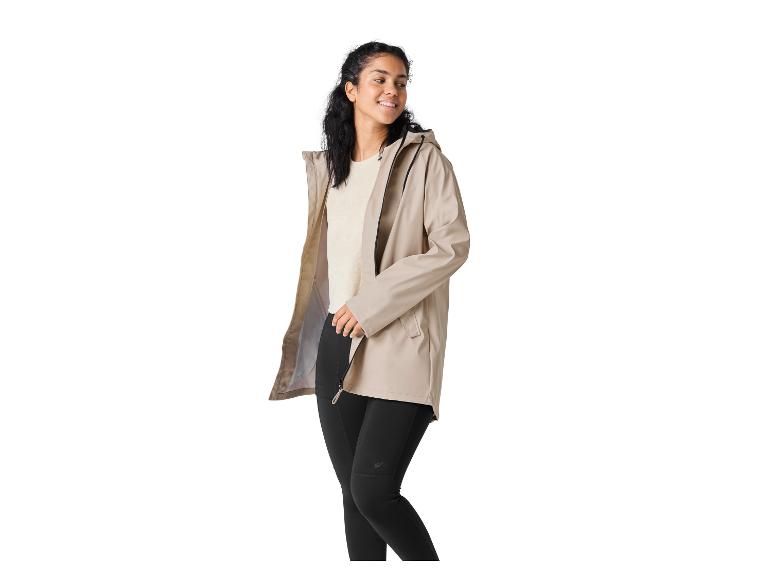 Femme en veste beige et leggings noirs, souriante et regardant vers la droite.