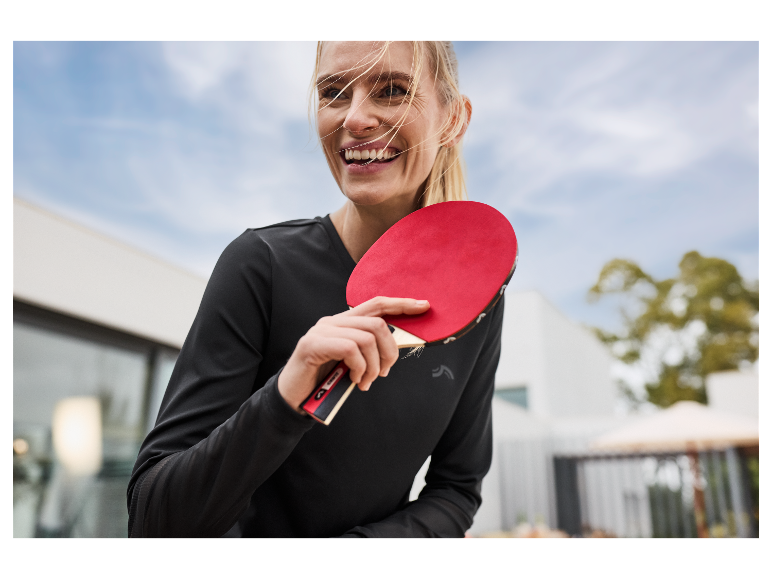 Une femme souriante tient une raquette de ping-pong rouge.