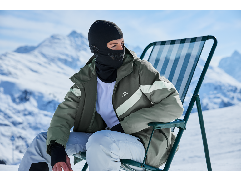Homme en cagoule et veste de ski verte assis sur une chaise dans les montagnes enneigées.