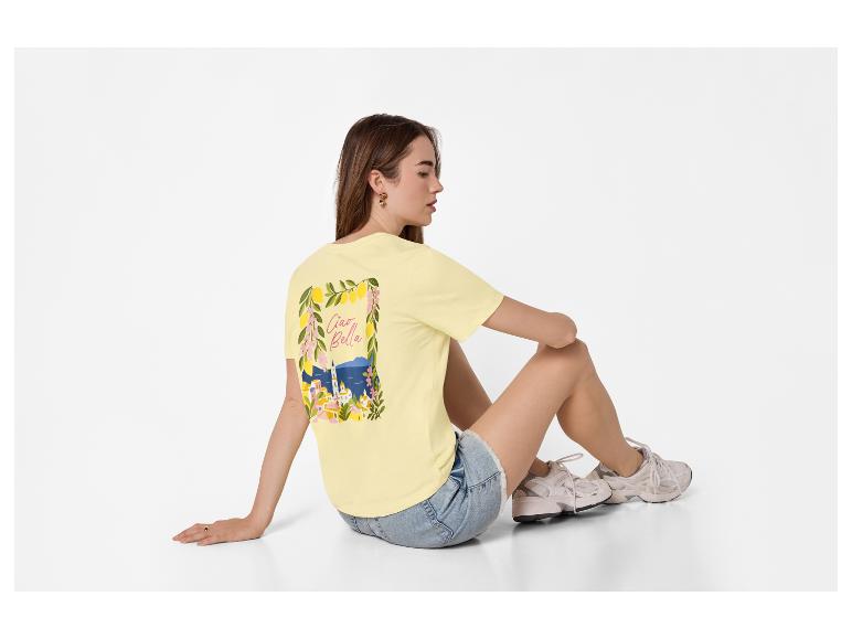 Femme en t-shirt jaune avec imprimé 'Ciao Bella' et short en jean, assise par terre.