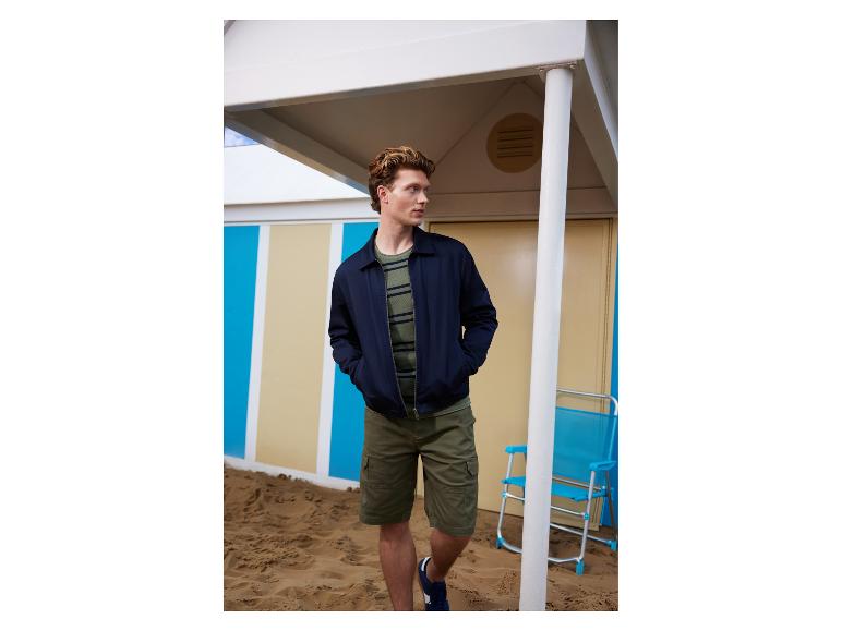 Homme en veste marine, t-shirt rayé et short cargo sur une plage de sable.