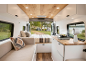 Intérieur de van avec cuisine, lit et canapé, vue sur un camping au bord du lac.