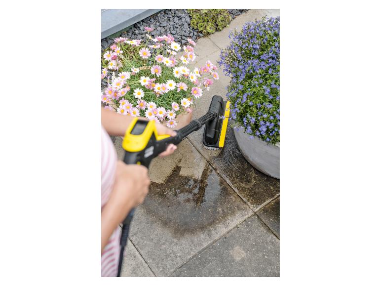 Femme nettoyant la terrasse avec un nettoyeur haute pression et une brosse, fleurs en arrière-plan.