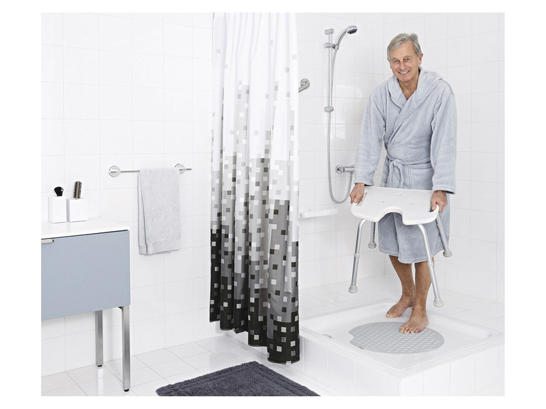 Un homme en peignoir se tient dans une salle de bain avec un tabouret de douche et un tapis antidérapant.