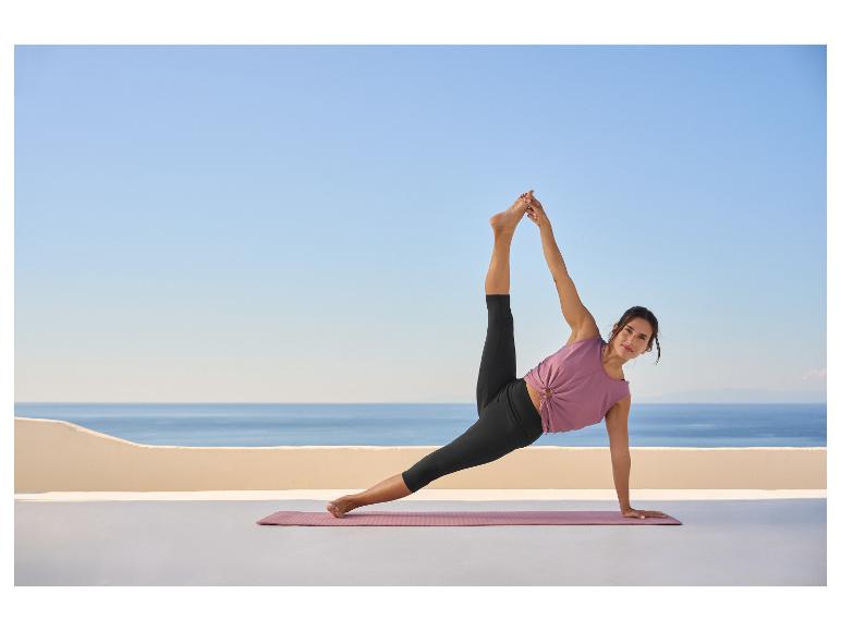Femme en haut violet et legging noir faisant du yoga sur un tapis sur une terrasse avec vue sur la mer.