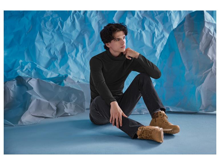 Homme en col roulé noir et jean assis par terre avec des bottes marron.