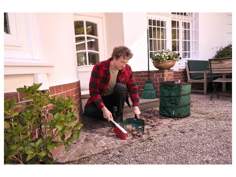 Homme balayant des feuilles avec un balai et une pelle dans un sac vert pour déchets de jardin.