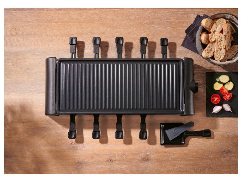 Appareil à raclette avec six poêlons, pain et légumes coupés sur une table en bois.