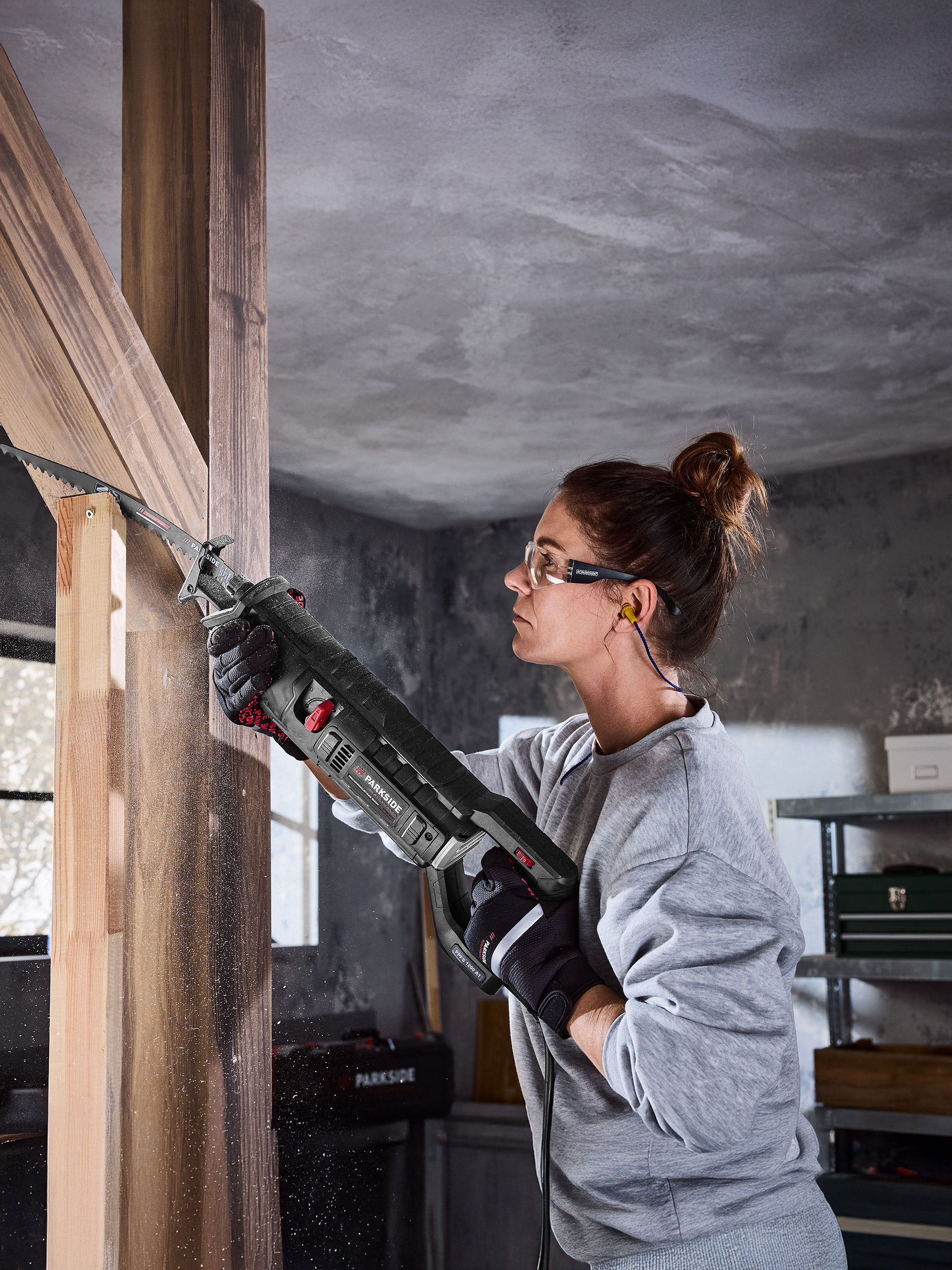 Femme utilisant une scie sabre Parkside pour couper des poutres en bois, avec lunettes de sécurité et protections auditives.