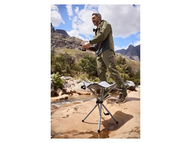 Homme pêchant dans une rivière de montagne, portant une veste et un pantalon kaki, avec un tabouret pliant.