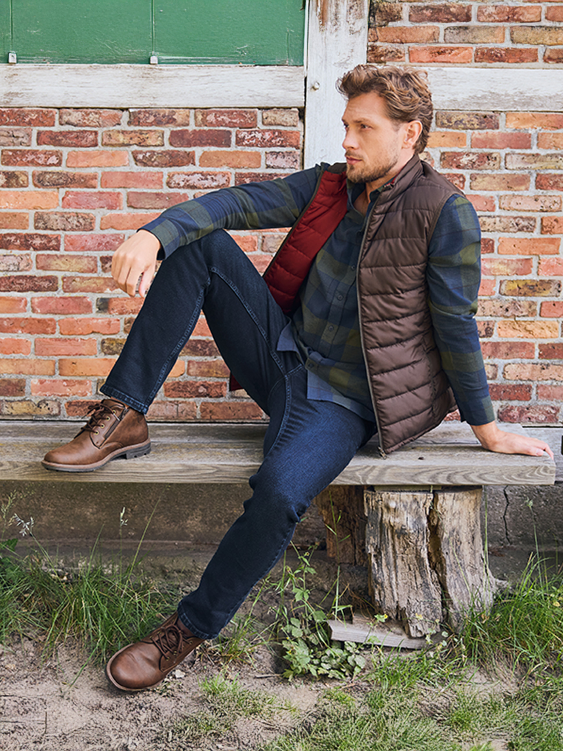 Homme en chemise à carreaux, doudoune sans manches et jean assis sur un banc en bois.