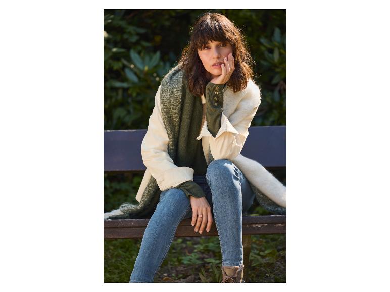 Femme en jean, pull vert et écharpe, assise sur un banc en bois.