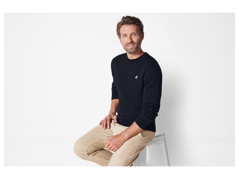 Un homme en pull noir en câble et en pantalon beige.