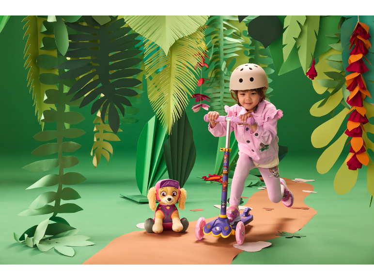 Fille avec un casque sur une trottinette, à côté d'un chien en peluche, dans un décor de jungle.
