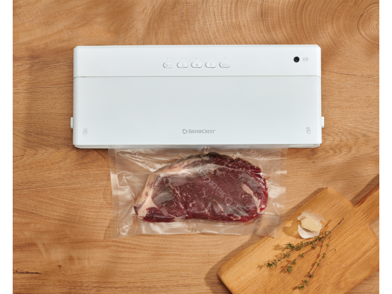 Machine sous vide avec bœuf scellé, ail et thym sur une planche à découper en bois.