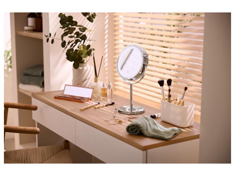 Coiffeuse avec miroir LED, pinceaux de maquillage et cosmétiques, avec plante et stores en arrière-plan.
