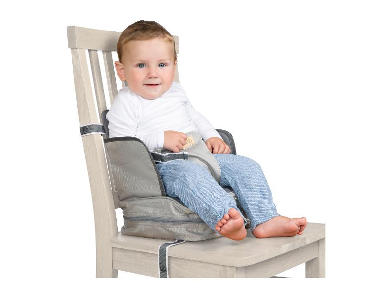 Un bébé souriant assis dans un rehausseur gris attaché à une chaise en bois.
